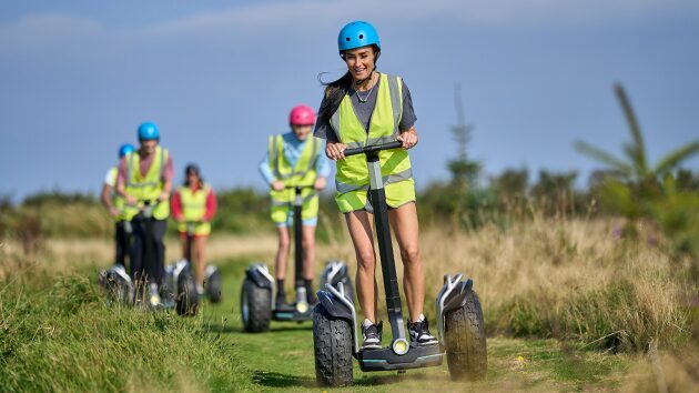 A diverse group of people wearing helmets and hi-vis jackets ride on the Segway Safari at Woolacombe Bay Holiday Park