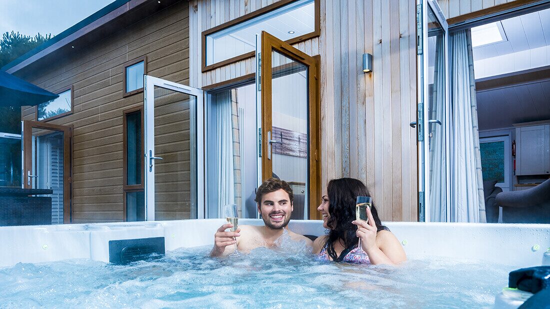 Couple soaking in a steaming outdoor hot tub at Surf Village lodges, relaxing with a drink in hand. A couple enjoys a steaming outdoor hot tub on a wooden deck by their lodge at Surf Village, holding glasses of wine or champagne as mist rises around them and coastal greenery frames the scene.