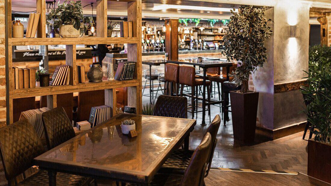 Stylish bar and restaurant interior with wooden tables, leather chairs, an open bookshelf with decorations, and a cozy atmosphere with warm lighting.