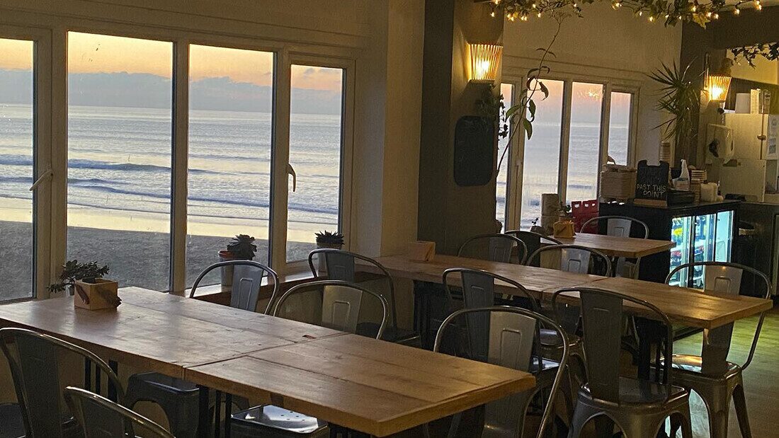 Beachcomber Café, Woolacombe Cosy beach café with wooden tables, indoor plants, and large windows offering stunning sunset views over the sea.