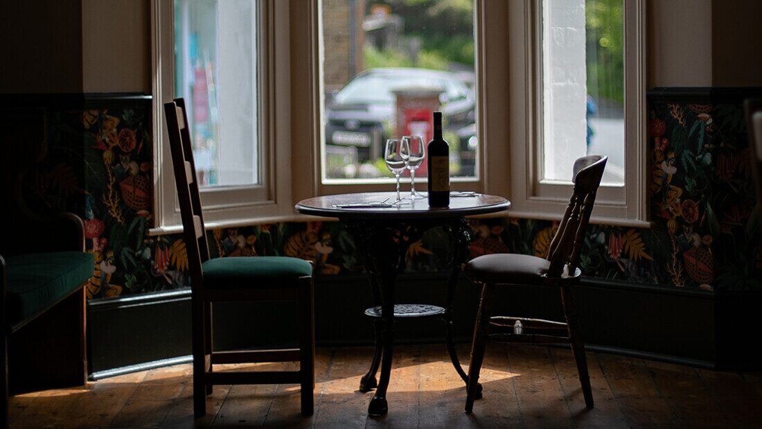 Mrs Fea's Bistro, Mortehoe Cosy bistro setting with two chairs by a round table, a bottle of wine, and glasses in front of bay windows.