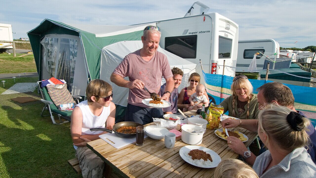 Family enjoying a meal together outside a caravan while camping, with food served at a picnic table
