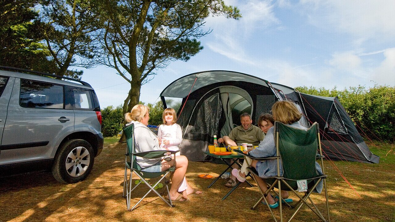 Family sitting outside a tent at a campsite, enjoying a meal together with car parked nearby