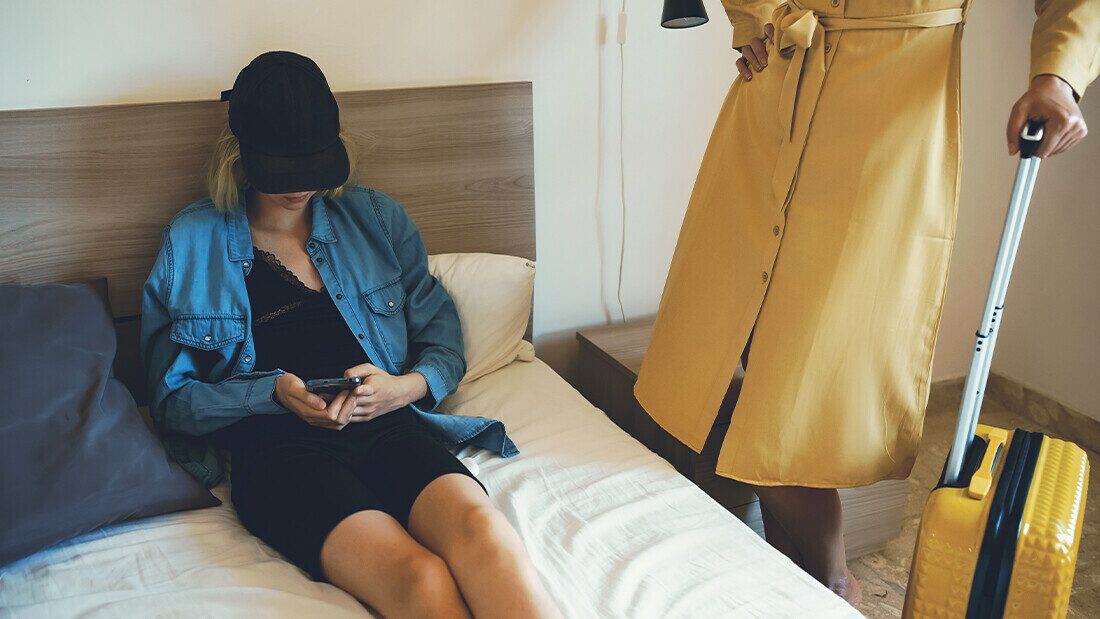 Budget hotels provide no activities or entertainment Teenager sitting bored on a hotel bed using a smartphone while a parent stands nearby with a suitcase.
