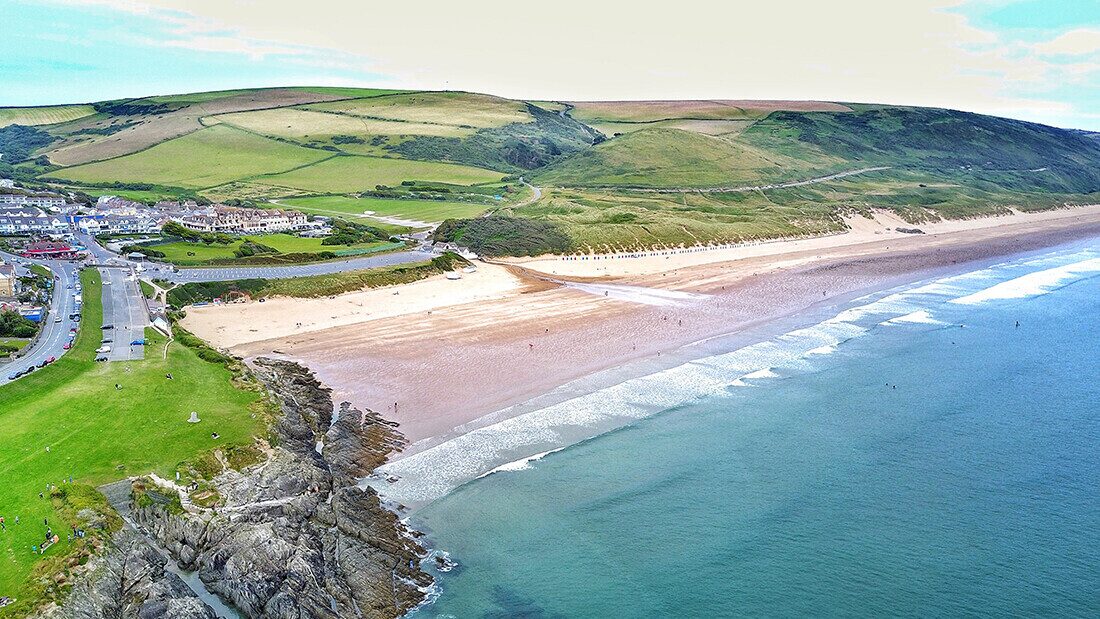 Aerial view of Woolacombe Beach in North Devon showing sandy shoreline, beach huts, car parks, and rolling green hills.