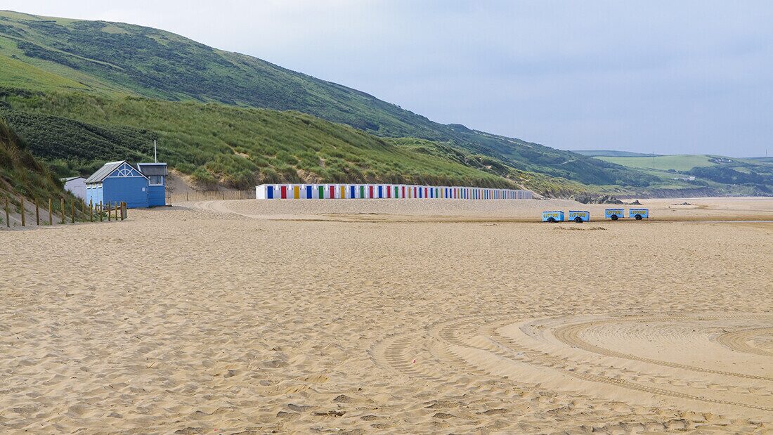 Row of colourful beach huts on Woolacombe Beach in North Devon with sandy shore and grassy dunes in the background.