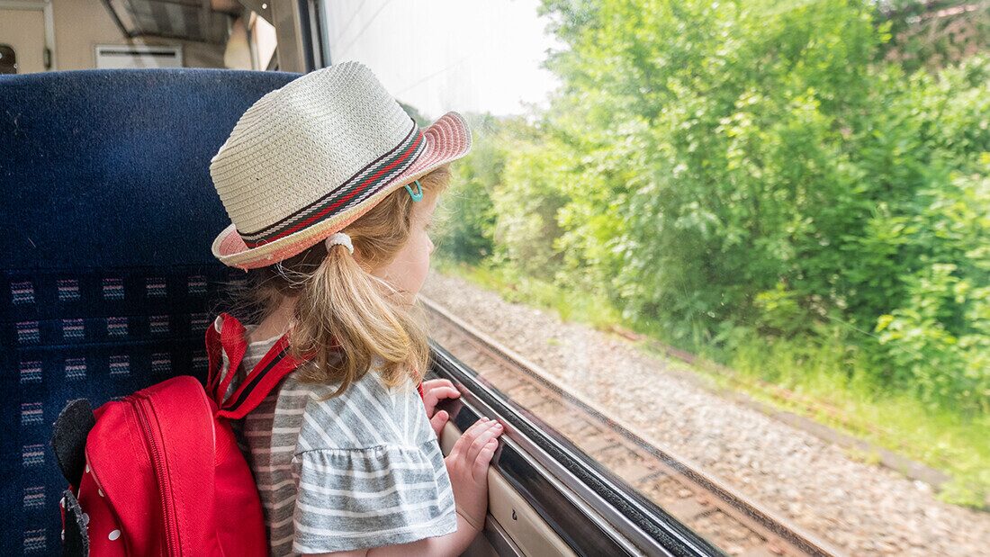 Little girl with red backpack looking out train window on summer family holiday journey through countryside.