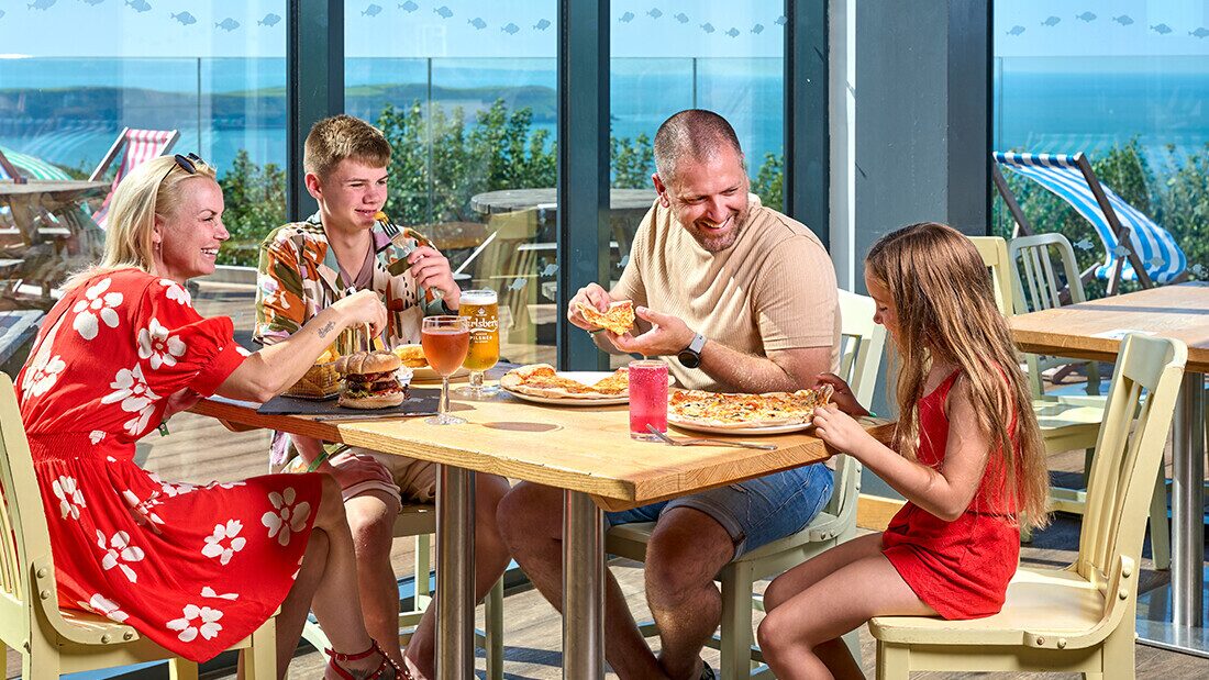 Family enjoying pizza and burgers together at a Woolacombe Bay Holiday Parks restaurant with sea views.