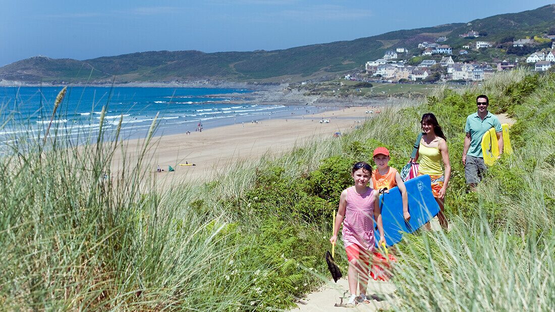 Bring everything you need for a beach day Family walking through sand dunes to Woolacombe Beach with bodyboards and beach gear on a summer holiday in North Devon.