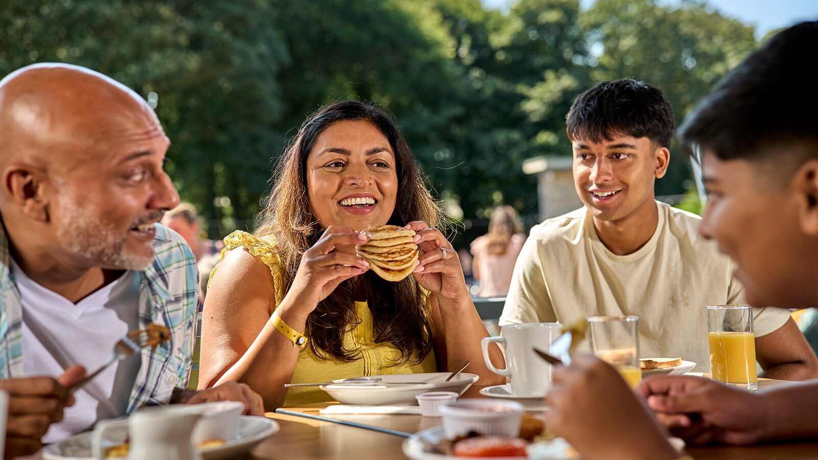 Woman smiling and eating a stack of American pancakes held in her hands whilst her family sits around her similing