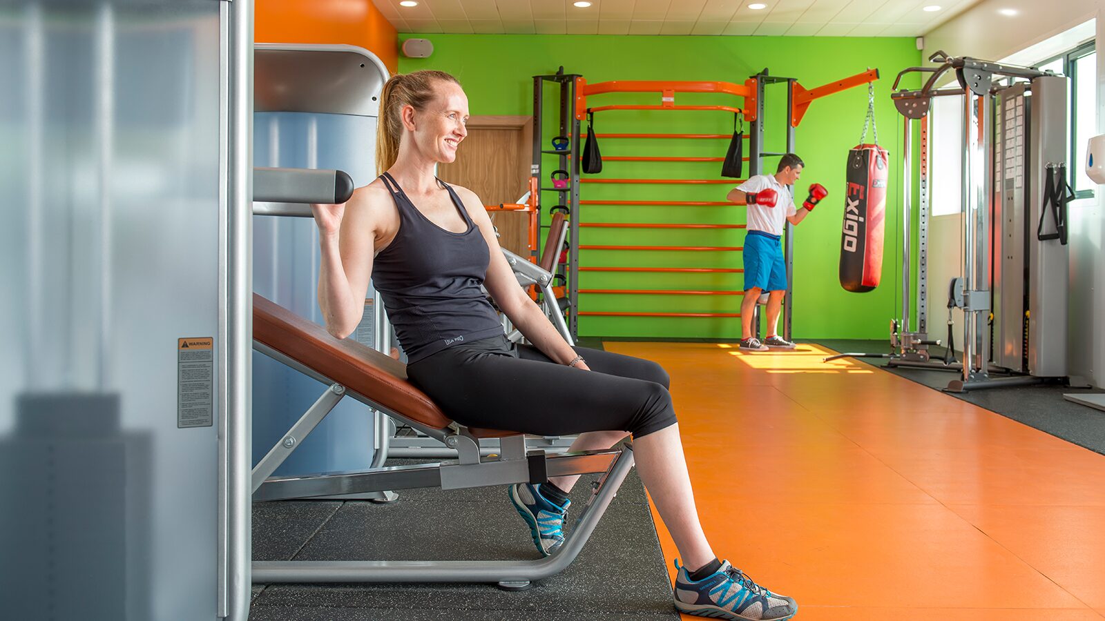A woman rests on a gym bench, surrounded by exercise equipment and a vibrant atmosphere whilst man wearing boxing gloves hits punchbag.