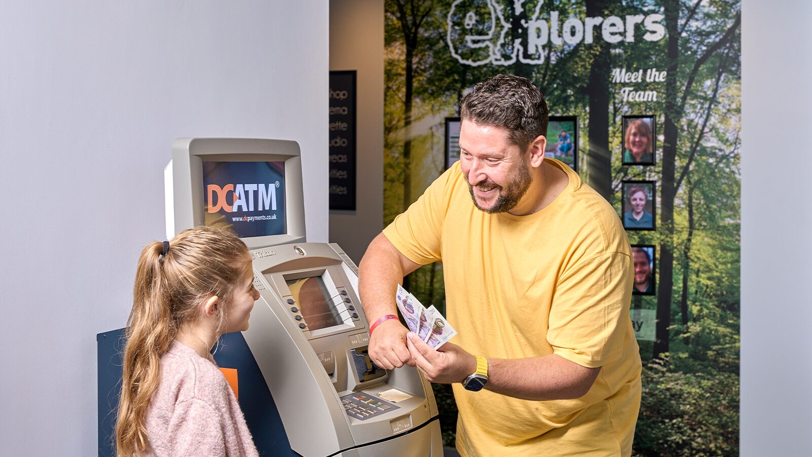 A man and a young girl stand together in front of an ATM machine, both looking at the cash.