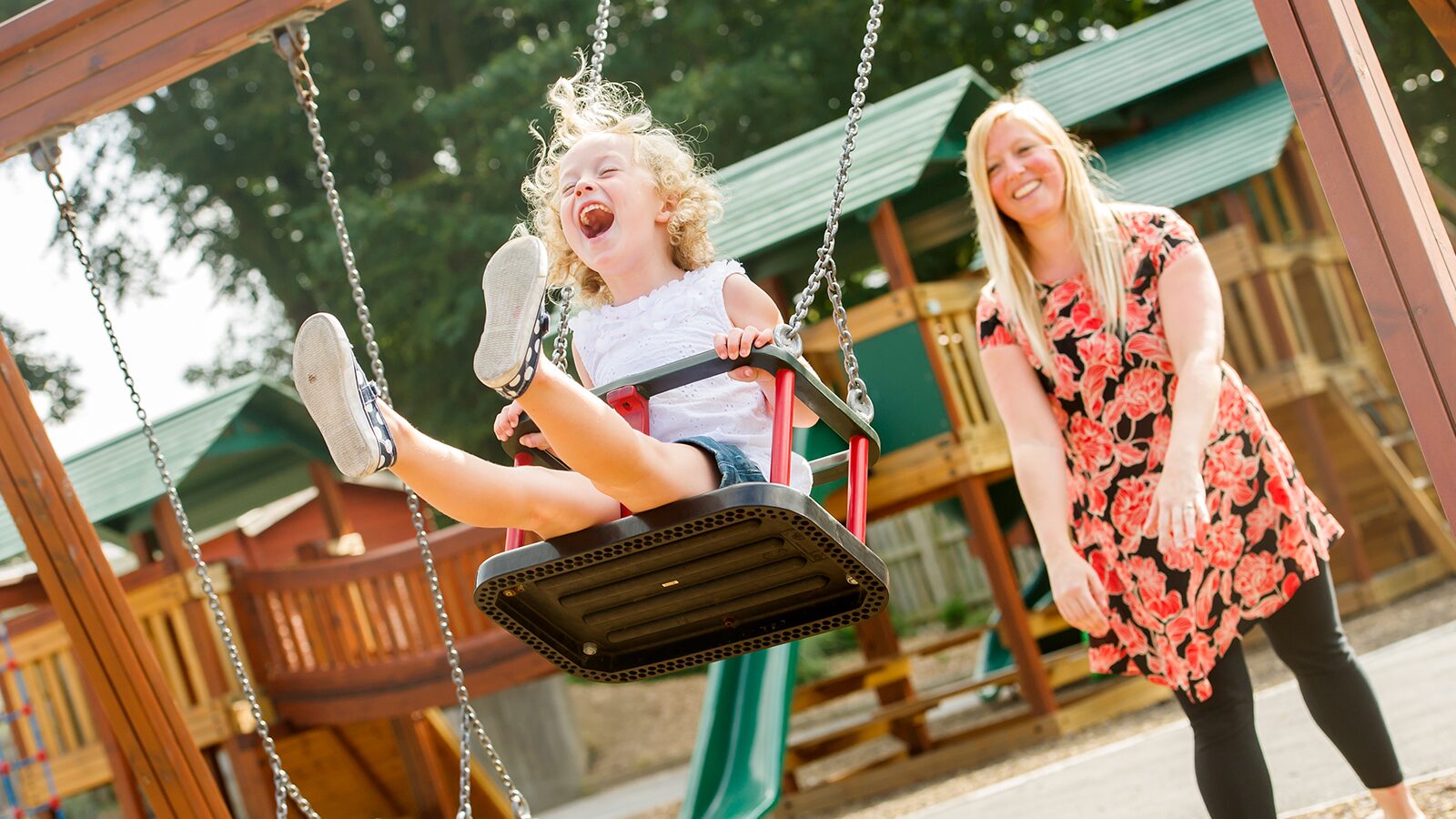 A woman and a child joyfully swinging together at a playground, surrounded by trees and colorful play equipment.