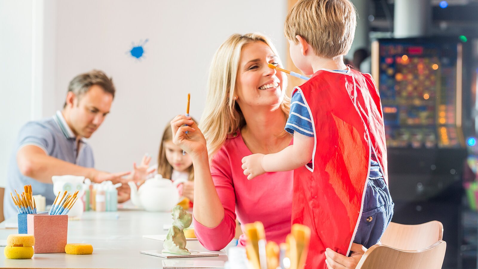 A young boy wearing a red apron tries to paint his mother whilst doing pottery painting at Woolacombe Bay Holiday Parks in Devon