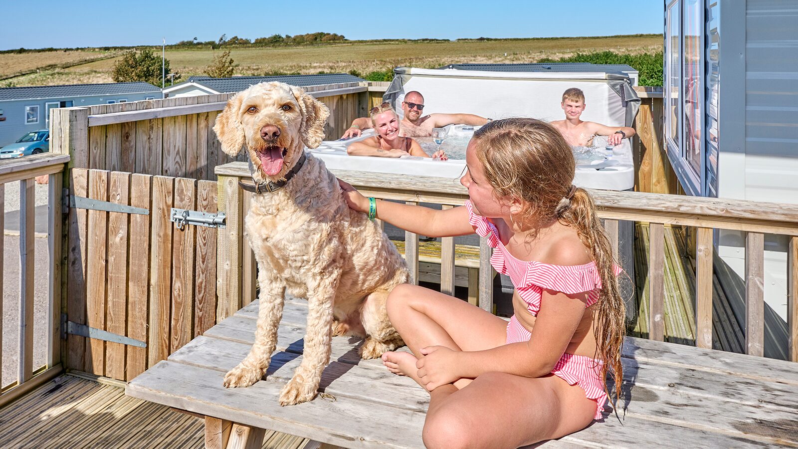 A family sits on a deck with their dog, next to a hot tub, enjoying a sunny day outdoors at Twitchen House Holiday Park.