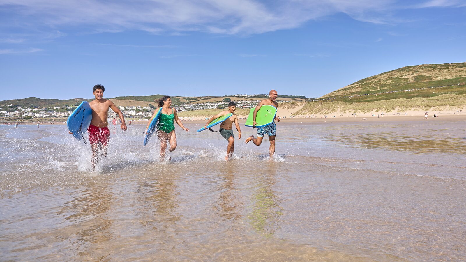 Family group holding body boards dashing into the sea at Woolacombe in North Devon, splashing water as they enter.