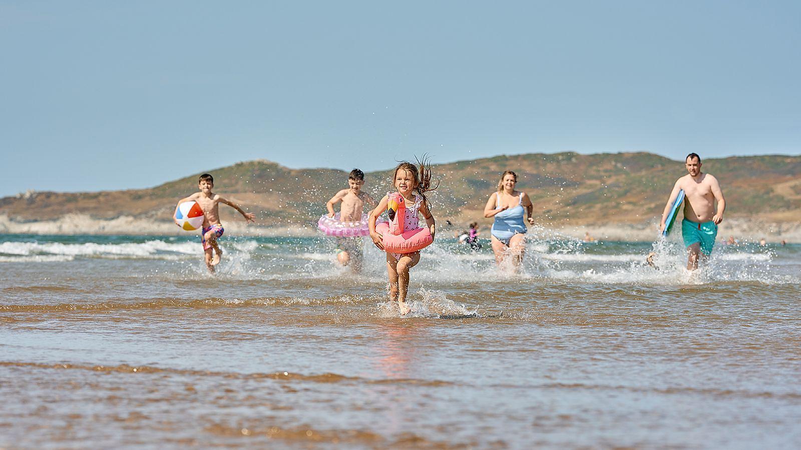 A young girl wearing a flamingo rubber ring running in the water at Woolacombe Sands Beach in North Devon with her family running behind her