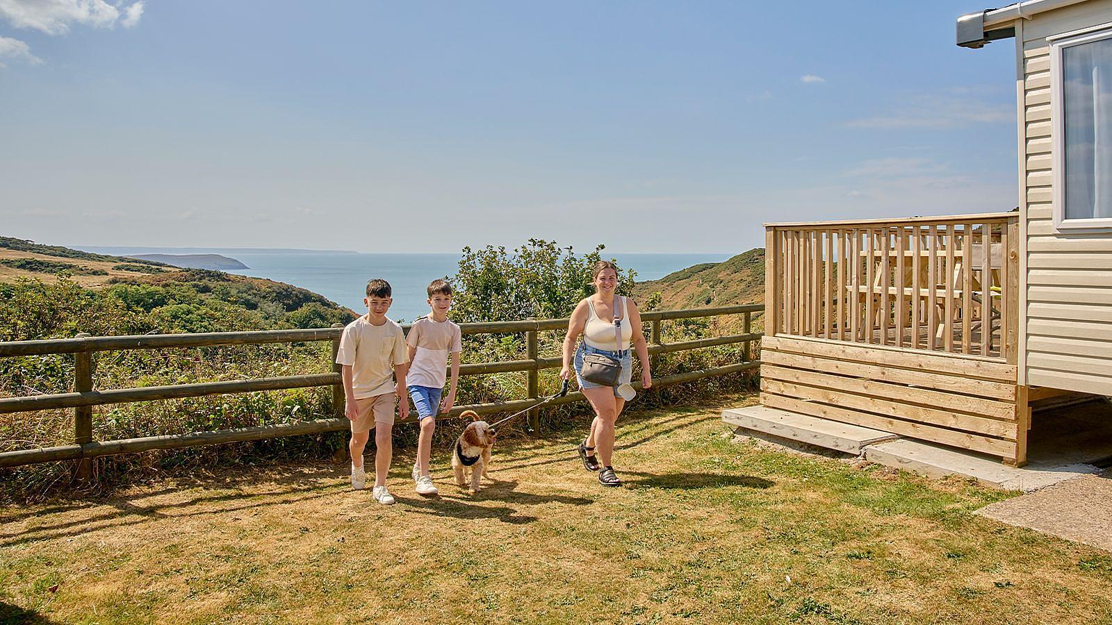 Outside a static caravan at Twitchen House Holiday Park in Woolacombe, three people and a dog gather, creating a cheerful scene with an Atlantic Ocean sea view backdrop.