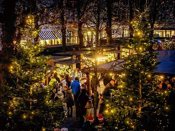 Gemütlicher Weihnachtsmarkt am Abend, mit Menschen, die zwischen beleuchteten Kiefern und stimmungsvollen Ständen unter warmem Licht spazieren.