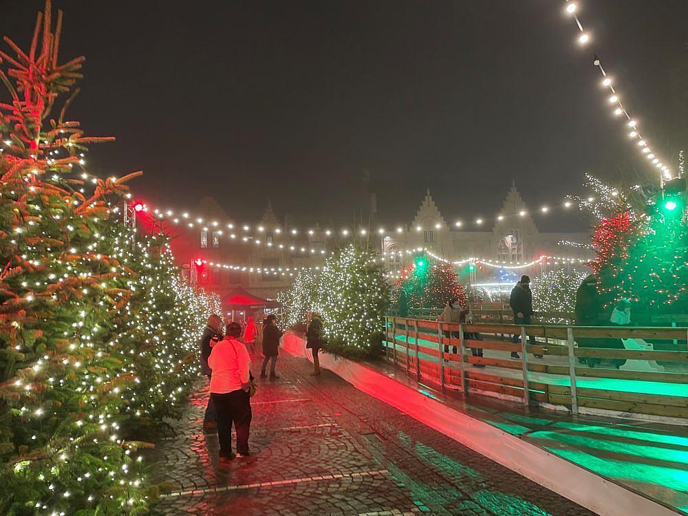 Stimmungsvoll beleuchtete Weihnachtsszene mit Menschen, die an hell erleuchteten Weihnachtsbäumen und einer Eisbahn entlang spazieren, unter Girlanden warmer Lichter an einem nebligen Abend.