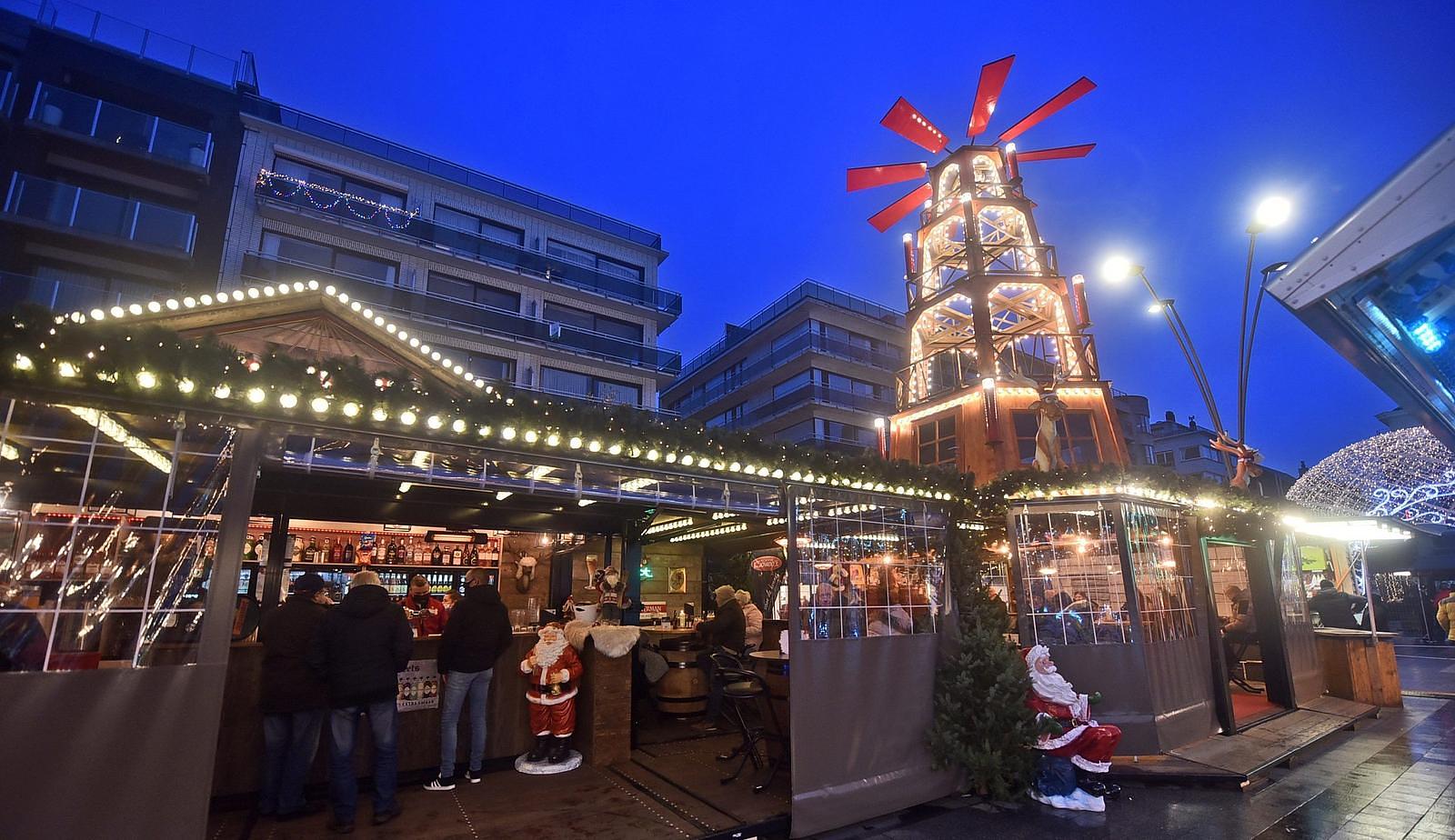 Gemütlicher Weihnachtsmarkt mit einem beleuchteten Holzturm, stimmungsvollen Ständen und Menschen, die an der Bar etwas trinken, vor der Kulisse moderner Gebäude am Abend.