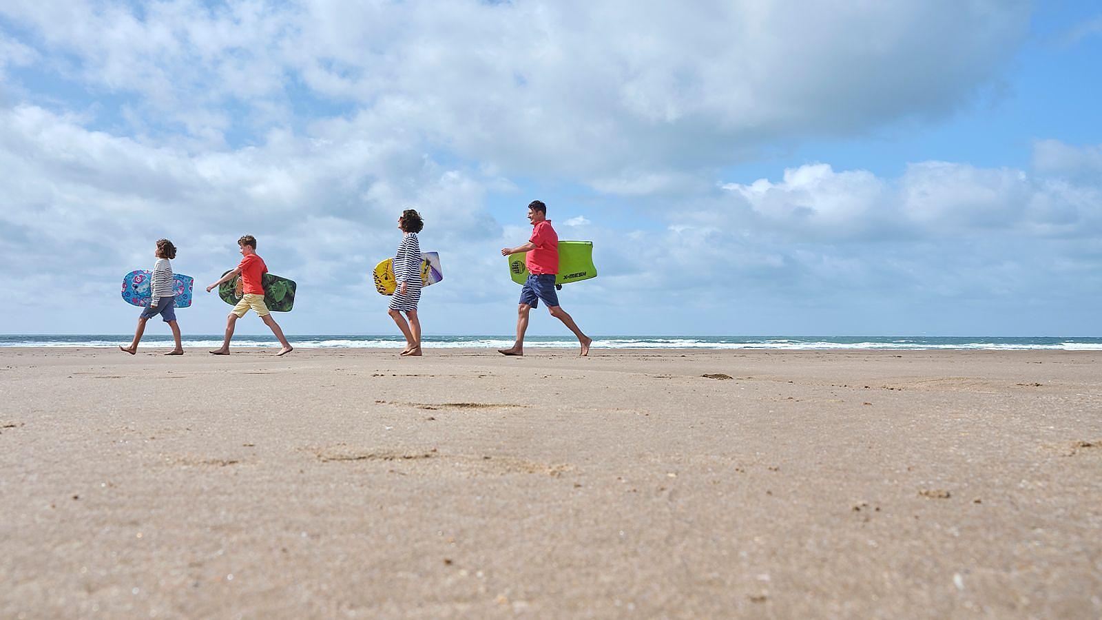 Family of four stroll on the sandy beach at Woolacombe in North Devon, holding surfboards, with the ocean waves gently crashing nearby.