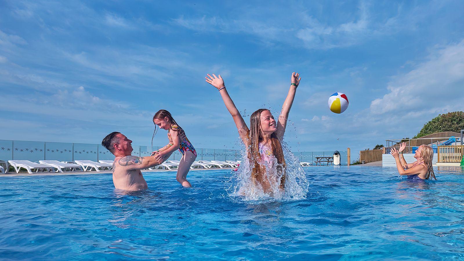 A family enjoys a sunny day playing with a ball in an outdoor swimming pool at Woolacombe Bay Holiday Park in North Devon, splashing and laughing together.