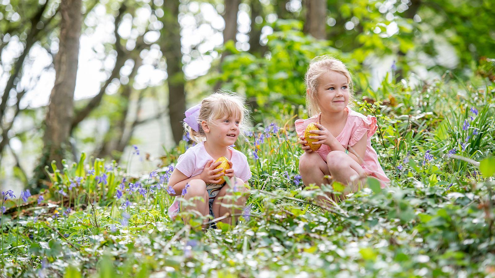 Two small girls sit on the grass at Twitchen House Holiday Park, holding golden coated chocolate Easter eggs and smiling at each other.