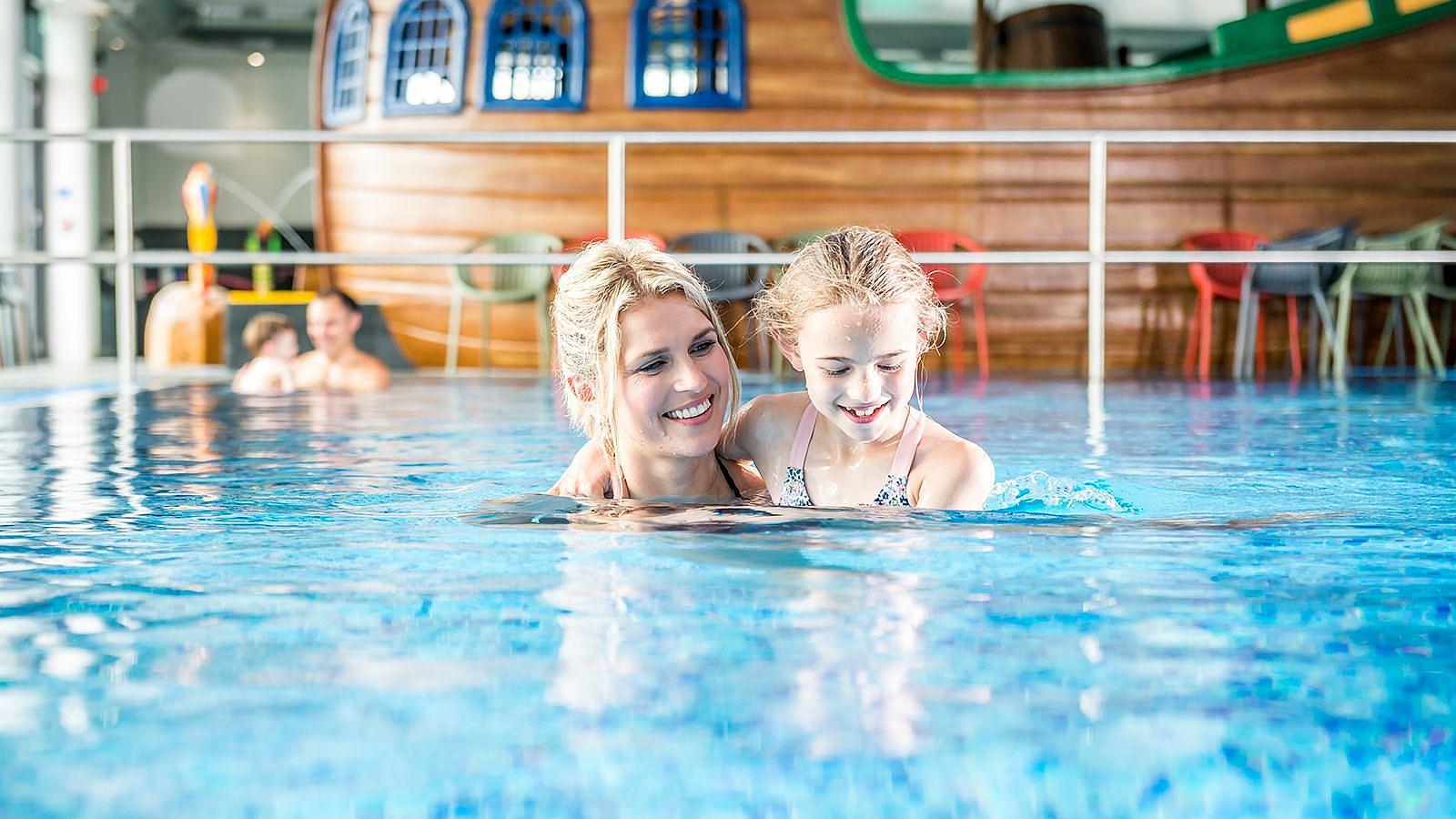 A woman and a child splash in an indoor pool at Woolacombe Bay Holiday Park, surrounded by bright sunlight and cheerful atmosphere.
