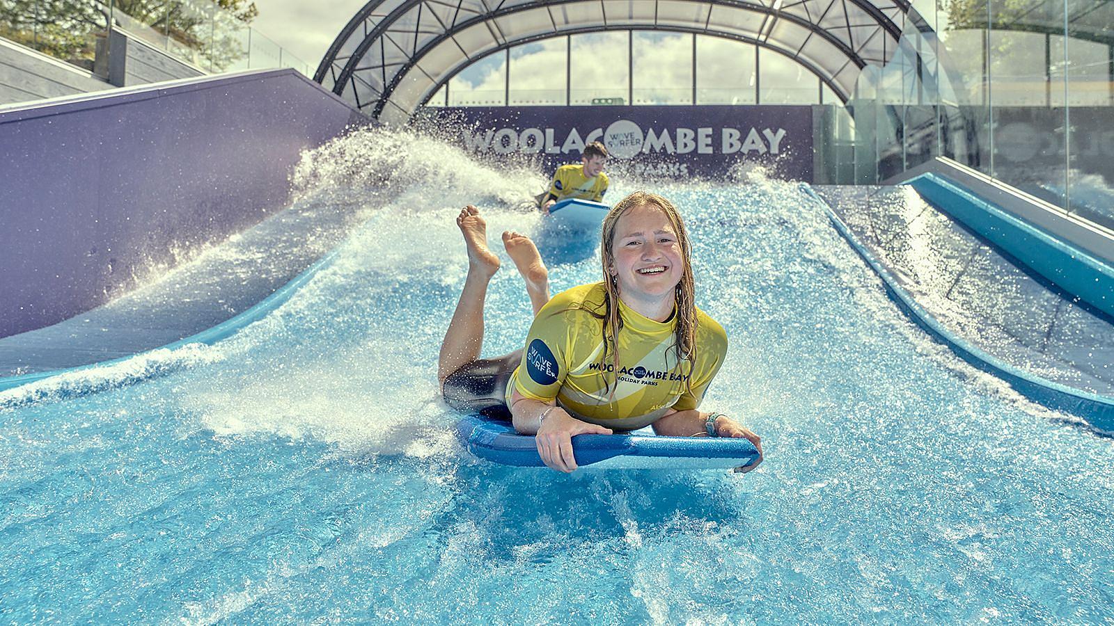 A young woman joyfully rides on the Wavesurfer surfing simulator at Twitchen House Holiday Park, splashing water around her.