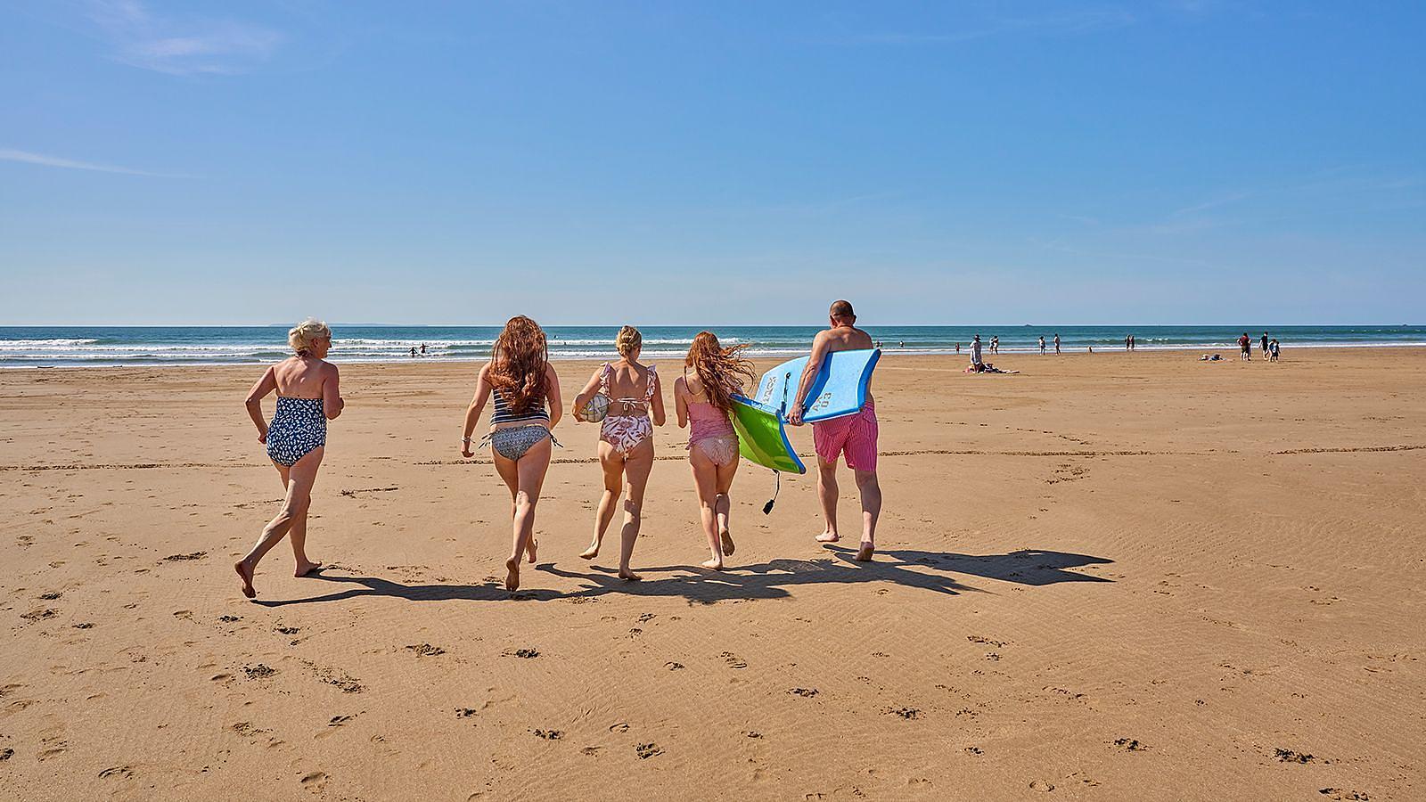 A diverse family group of people running on Woolacombe Sands beach some holding body boards, surrounded by sand and the gentle surf of the ocean.