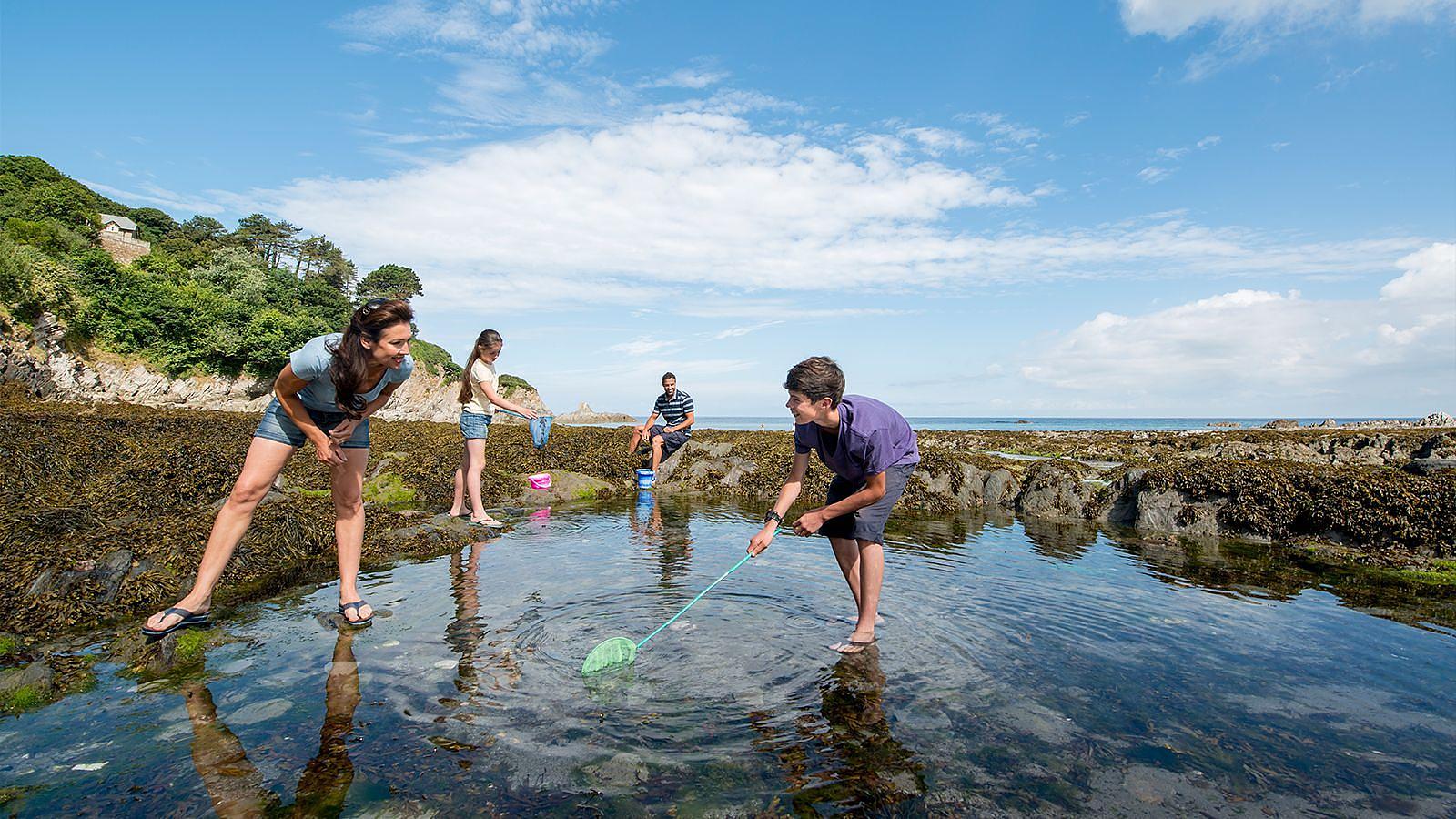 A family group gathered in shallow water at Lee Bay in North Devon, engaging in rockpooling leisure activities on a warm spring day.
