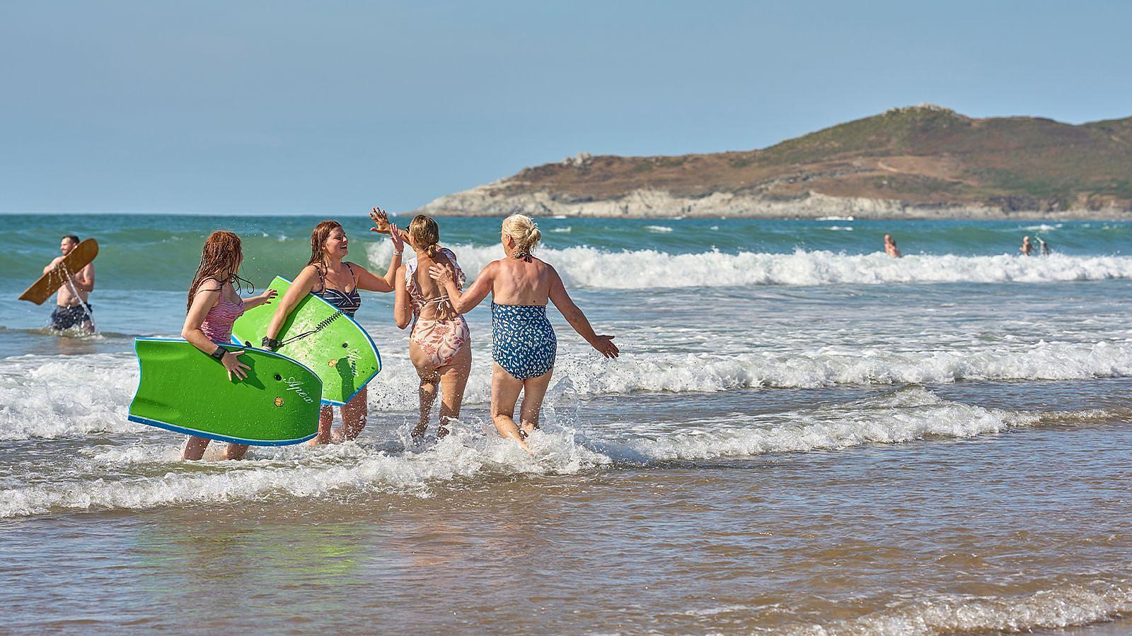 Several women with surf body boards stride into the ocean at Woolacombe, preparing for a fun day of surfing and enjoying the waves.