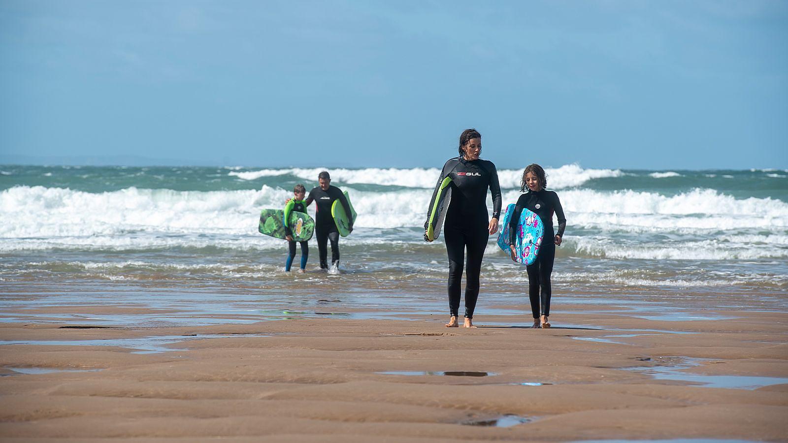 A family of beachgoers walks on the sand on Woolacombe Beach, each with a surfboard ready for surfing and body boarding