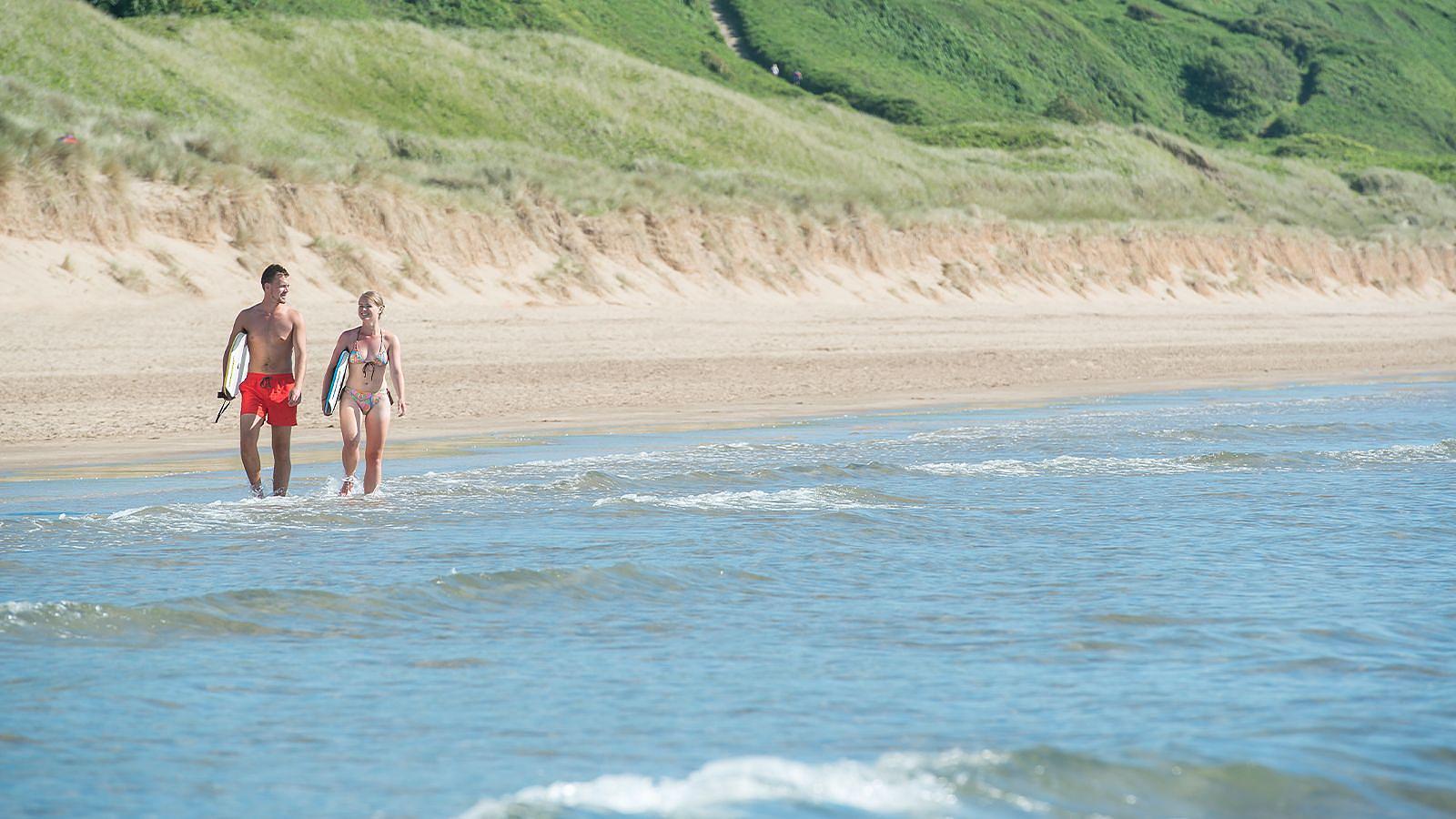 A young couple walking along the sandy beach in Woolacombe holidng body boards ready to go into the sea