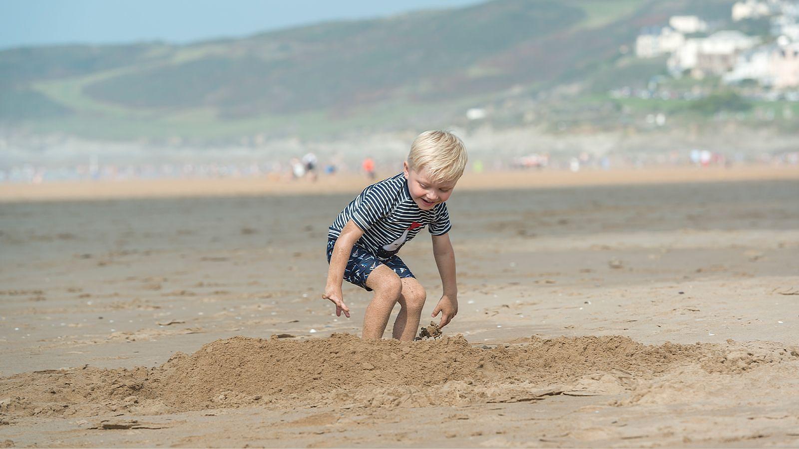 A young boy joyfully playing in the sand on a sunny beach at Woolacombe in North Devon, surrounded by gentle waves and a clear blue sky.