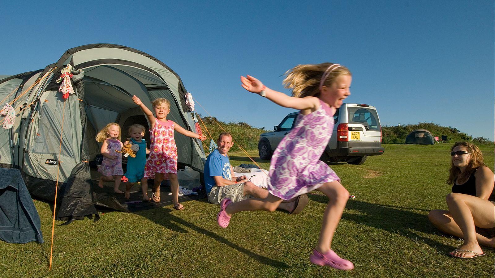 A young girl joyfully jumps in the air, surrounded by her smiling family in a sunny outdoor setting when camping at Twitchen House Holiday Park in Woolacombe