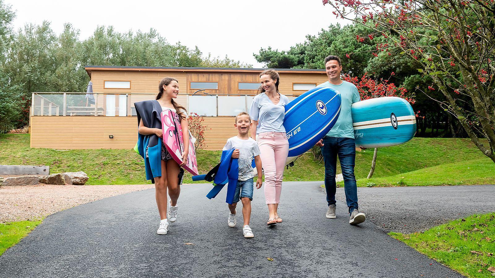 A family strolls along the road in Surf Village at Woolacombe Bay Holiday Park in North Devon, surfboards in hand, excited for their beach outing.