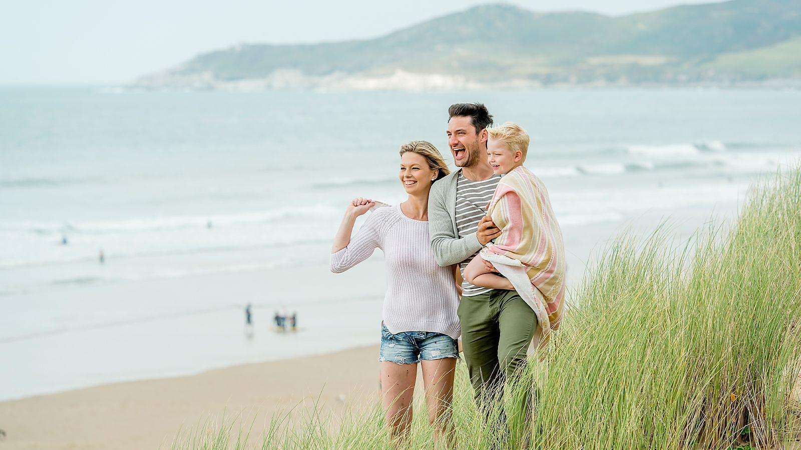 A man and woman walk along the sand dunes at Woolacombe beach with their child, enjoying an Autumn day by the ocean.