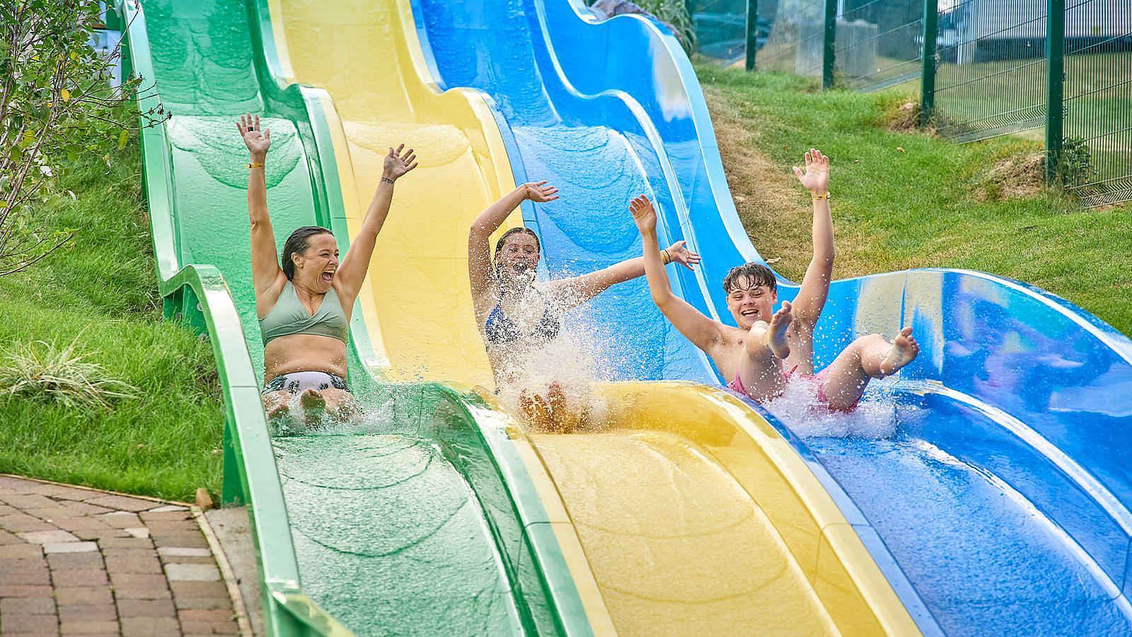 Three excited people sliding down a large water slide at a lively water park at Twitchen House in Woolacombe, splashing water around them