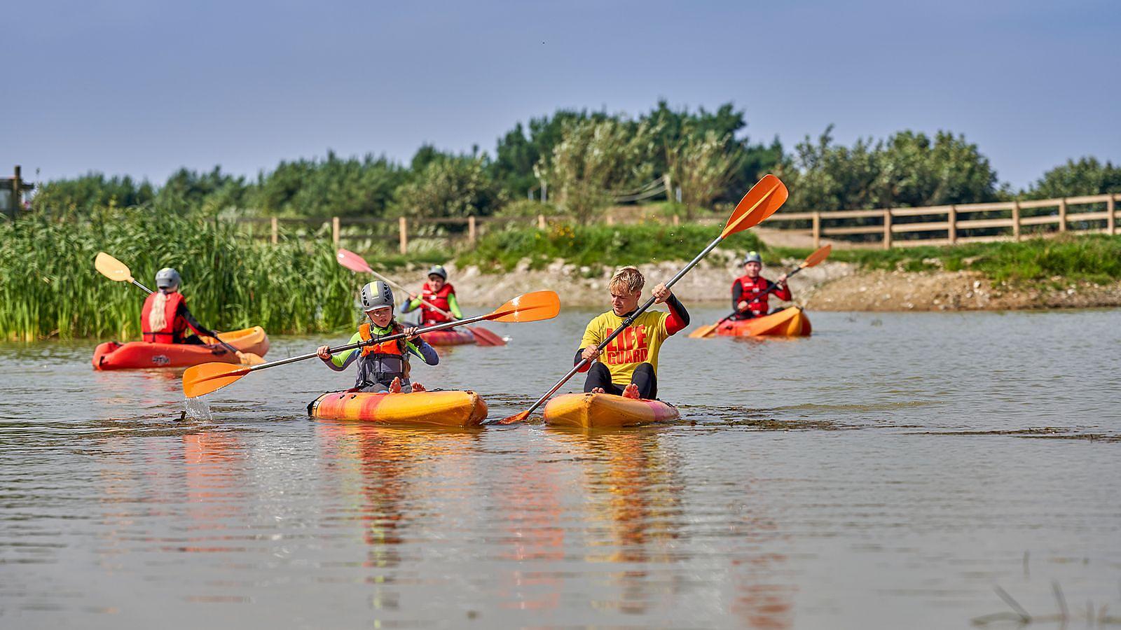 Several individuals in colourful kayaks enjoying a sunny day on a calm lake, with lush greenery nearby at Woolacombe Bay Holiday Park in North Devon.