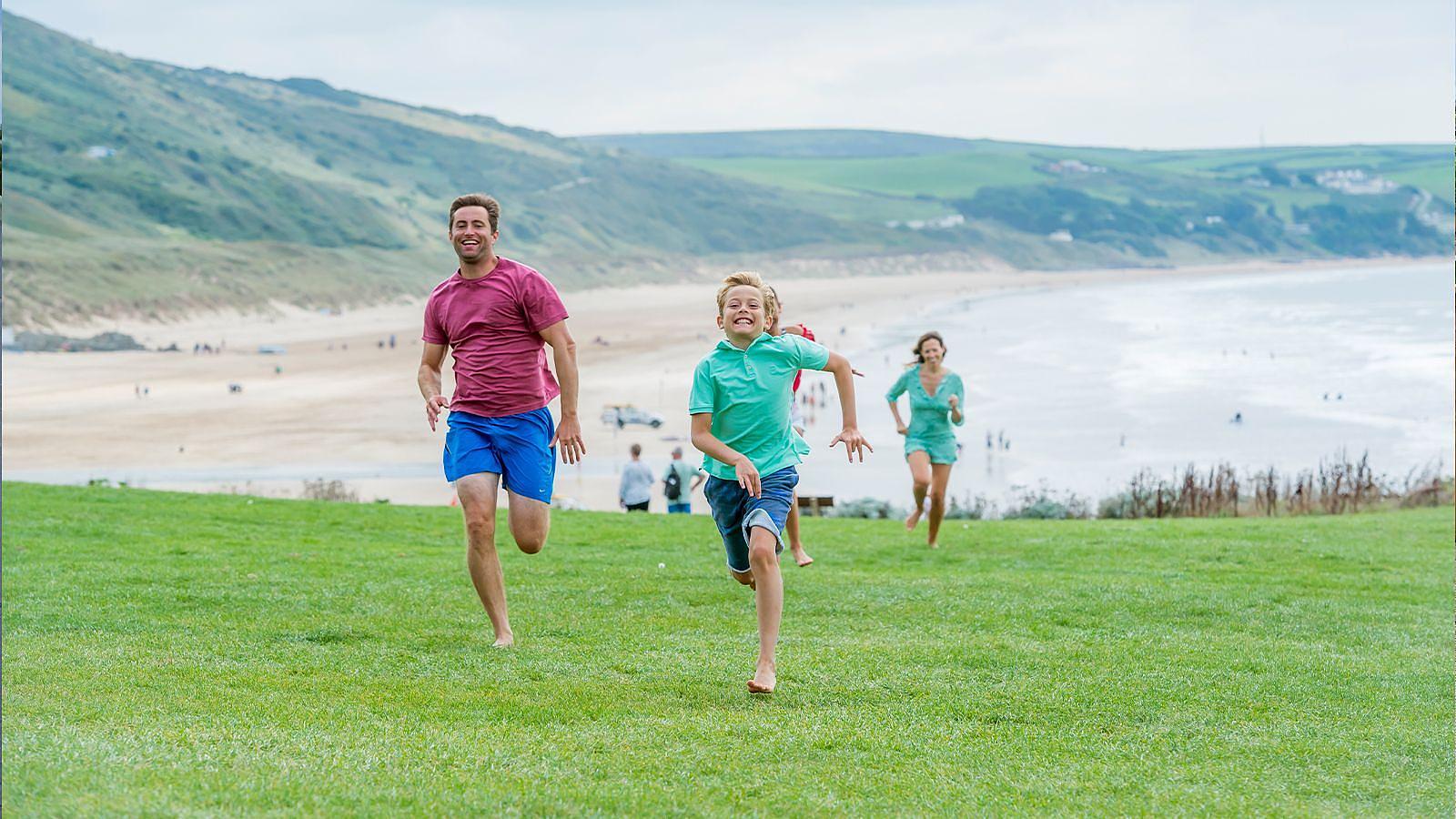 A family of four sprinting along the shoreline in North Devon, enjoying a sunny day at the beach at Woolacombe with the ocean in view.