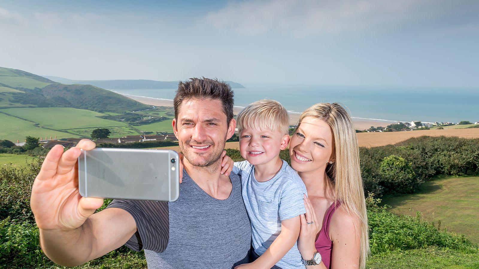 A man and woman smiling as they take a selfie with their young son, capturing a joyful family moment together at Woolacombe Bay Holiday Parks in Devon.
