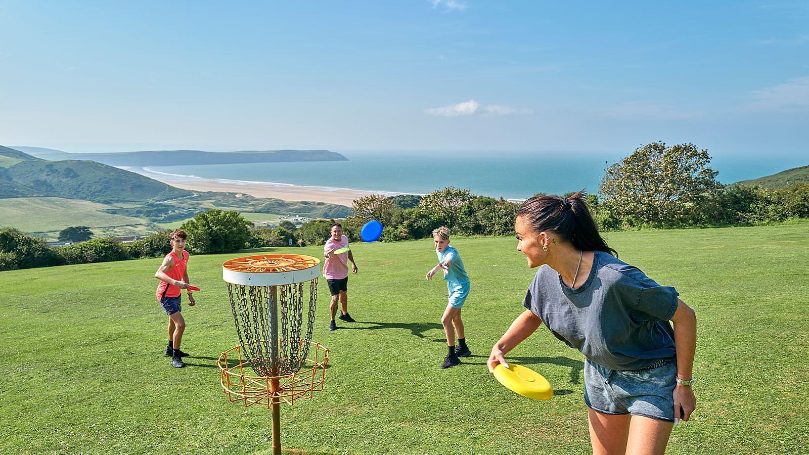 A family group enjoying a game of frisbee on a sunny grassy field at Woolacombe Bay Holiday Park with the beach and ocean behind