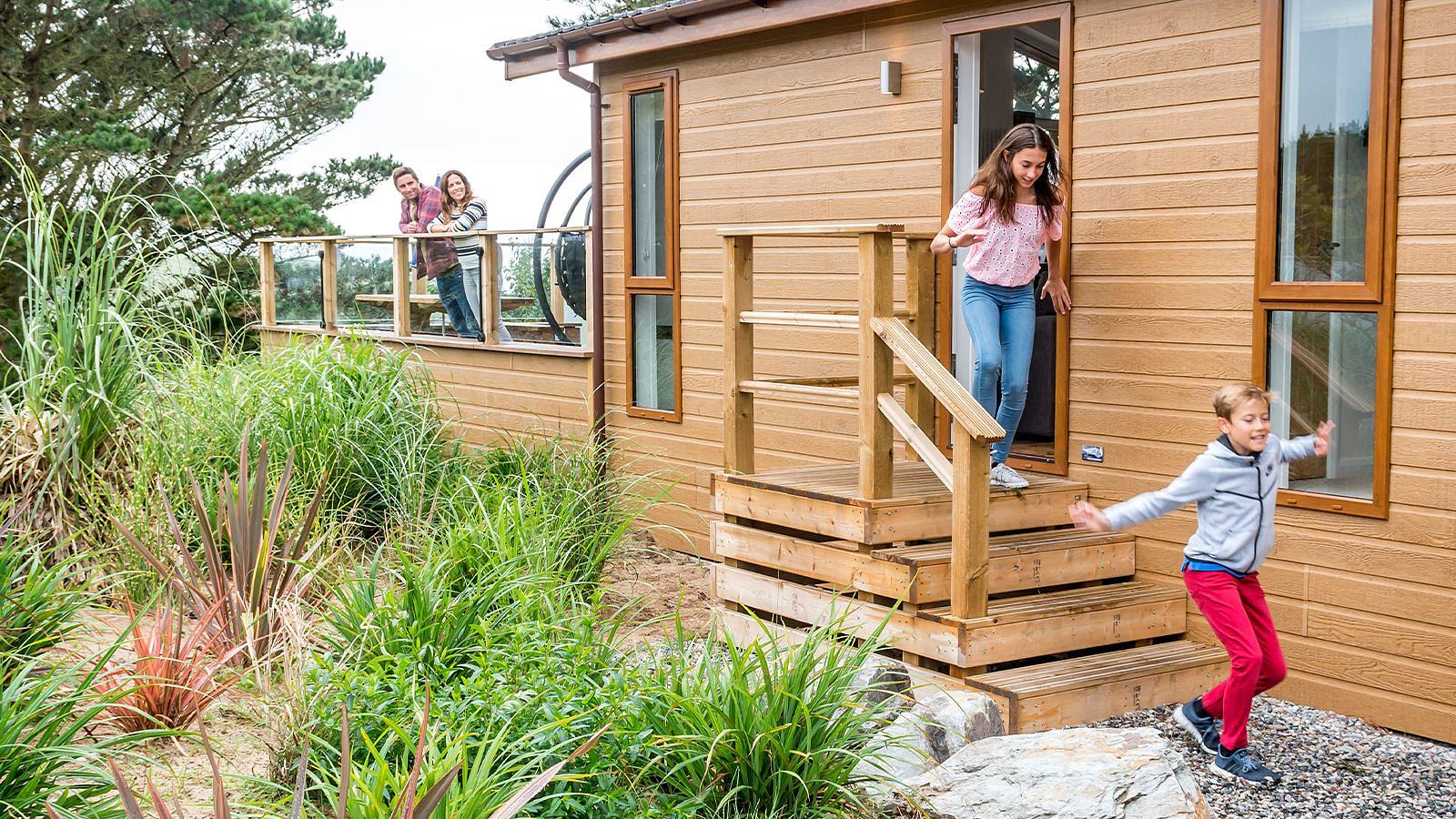 A family poses in front of a cozy lodge cabin, surrounded by trees and enjoying a sunny day outdoors at Woolacombe Bay Holiday Park.