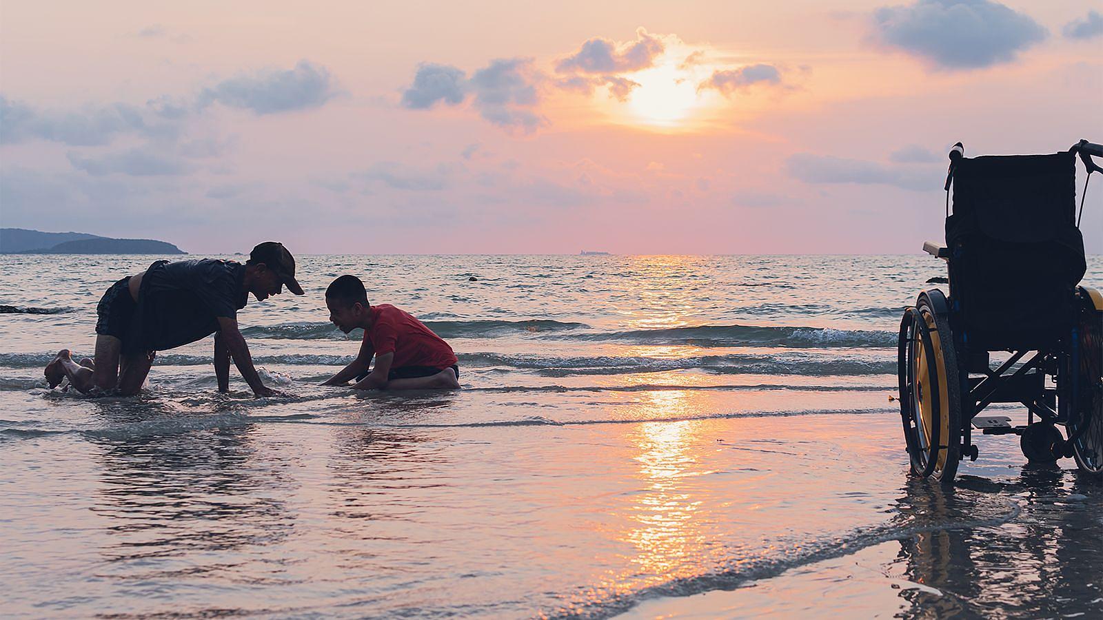 A man and a child having fun playing on the beach, with the ocean in the background and sunlight shining down whilst a wheelchair rests aside.