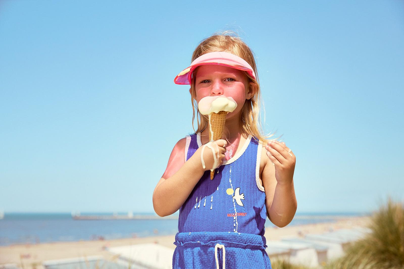 Enfant avec des lunettes de soleil roses et une casquette mange une glace en étant sur la plage, avec la mer en arrière-plan.