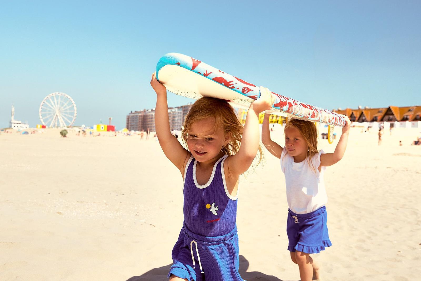 Two children are playing on the beach and carrying a surfboard together, with the sea and the Ferris wheel in the background.