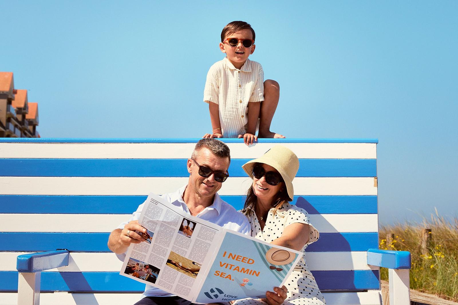Eine Familie genießt einen sonnigen Tag am Meer in Westende: Die Eltern sitzen auf einer bekannten blau-weiß gestreiften Küstenbank, während sie gemeinsam eine Zeitschrift lesen, und ihr Kind sitzt oben auf der Bank mit Sonnenbrille. Im Hintergrund sind Dünen und ein klarer blauer Himmel zu sehen.