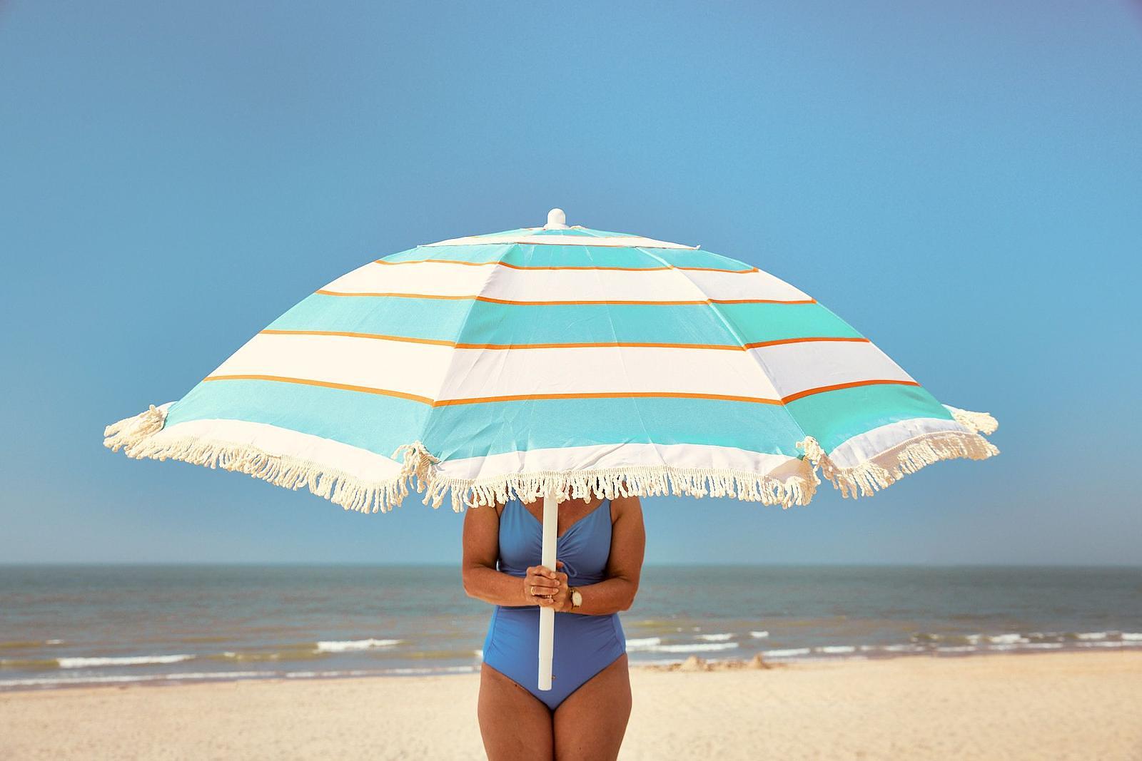 Une personne sur la plage tient un grand parasol de plage rayé au-dessus de sa tête, avec la mer en arrière-plan.